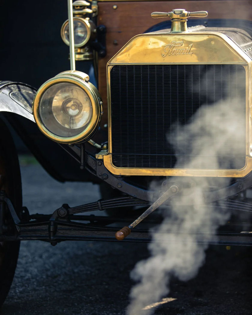 The brass radiator and crank handle of an unmarked vintage car under warm side light against a cool dark ground, a puff of exhaust drifting past
