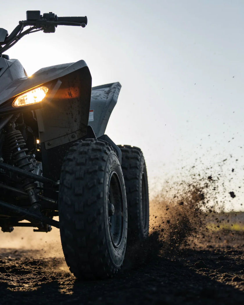A plain unmarked quad bike cornering hard under warm low sun against a cool dark ground, mud flying from the rear tyres