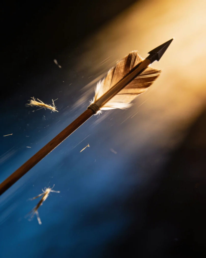 A wooden arrow caught mid-flight in warm side light against a cold dark ground, its feathered fletching blurred with speed