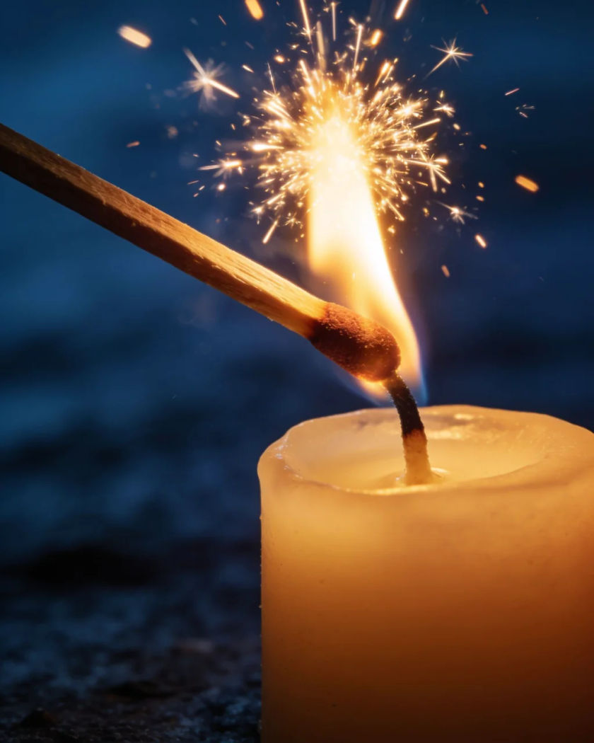 A match flaring as it touches a candle wick under warm light against a cold dark ground, tiny sparks lifting off the head