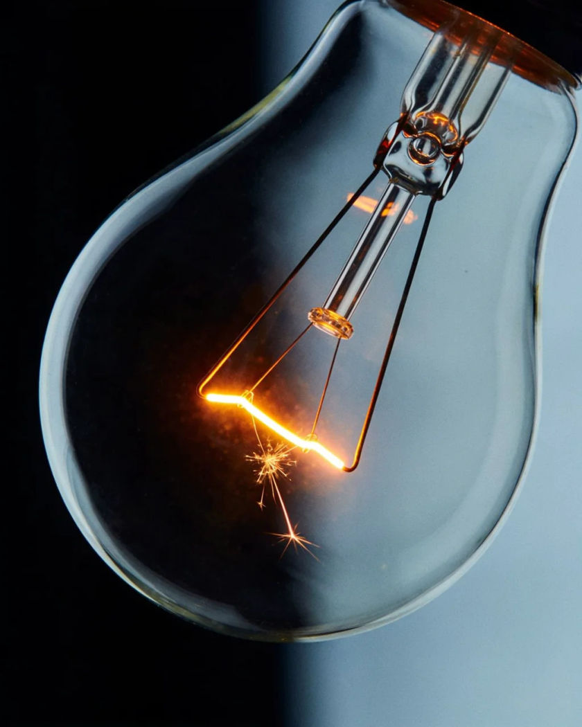 A bare clear glass light bulb hanging at an angle, its filament flaring bright orange with a tiny spark inside against a cold dark ground