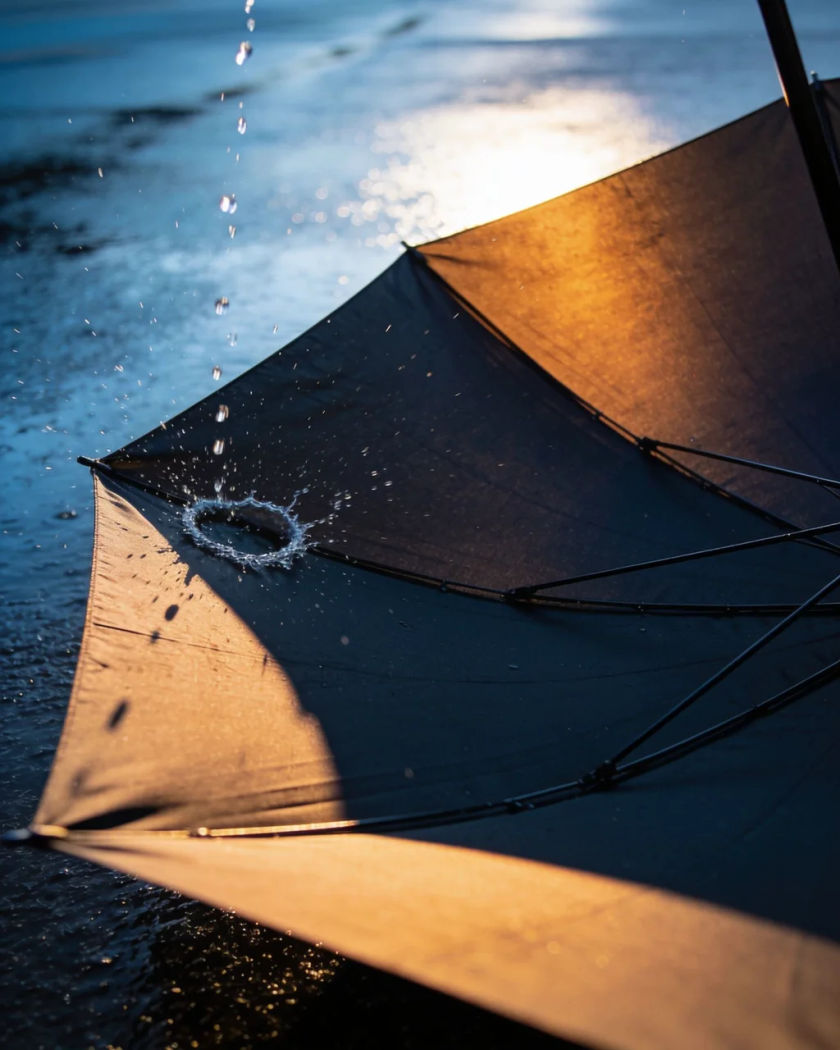 A dark umbrella canopy snapping open under warm side light against a cool dark ground, water droplets flung from its edge