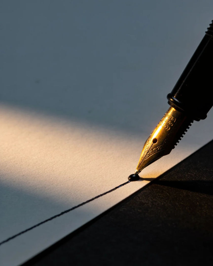 A plain fountain pen nib pressed to a blank sheet of paper under warm side light against a cold dark ground, a bead of wet ink glinting at the tip