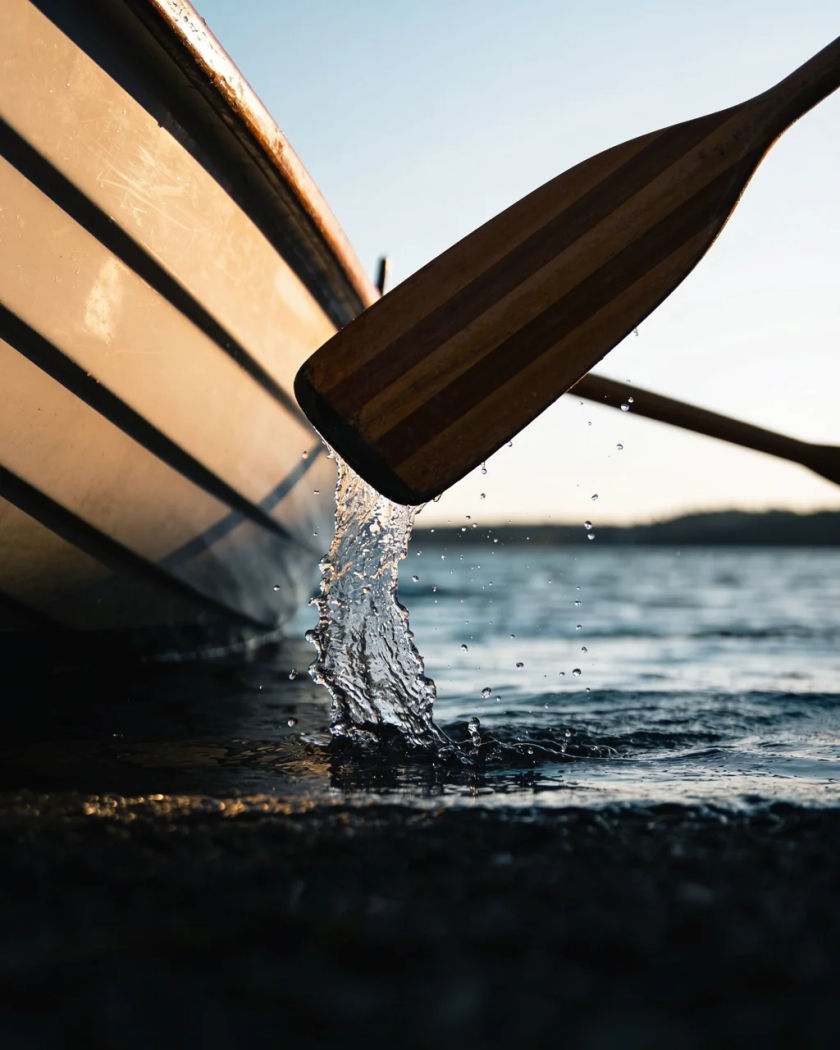 A wooden oar blade lifting out of dark water beside a rowboat hull under warm side light against a cold dark ground, drops spraying from the blade
