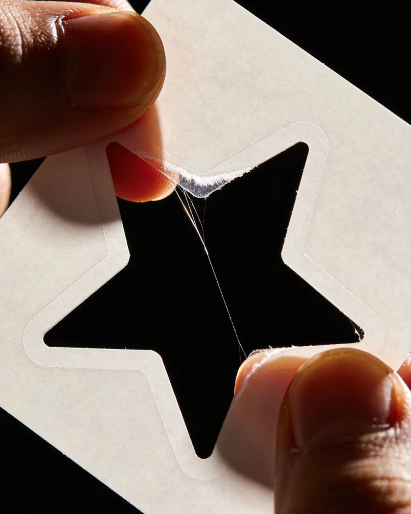 A glossy star-shaped sticker peeling away from its backing sheet between two fingers under warm side light against a cool dark ground, the glue stretching at the edge