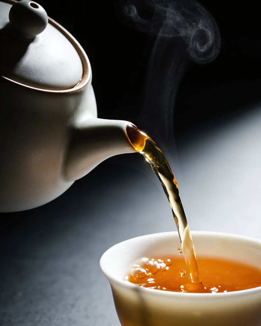 A plain ceramic teapot pouring a glowing amber stream of tea into a white porcelain cup under warm side light against a cool dark ground, steam curling and droplets splashing at the rim