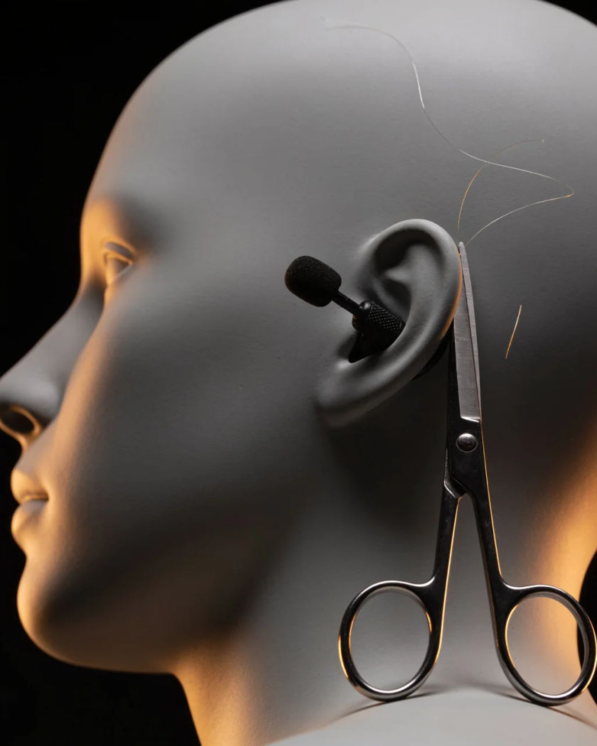 A pair of steel scissors open beside the ear of a plain dummy head under warm side light against a black ground, a few snipped threads drifting in the air