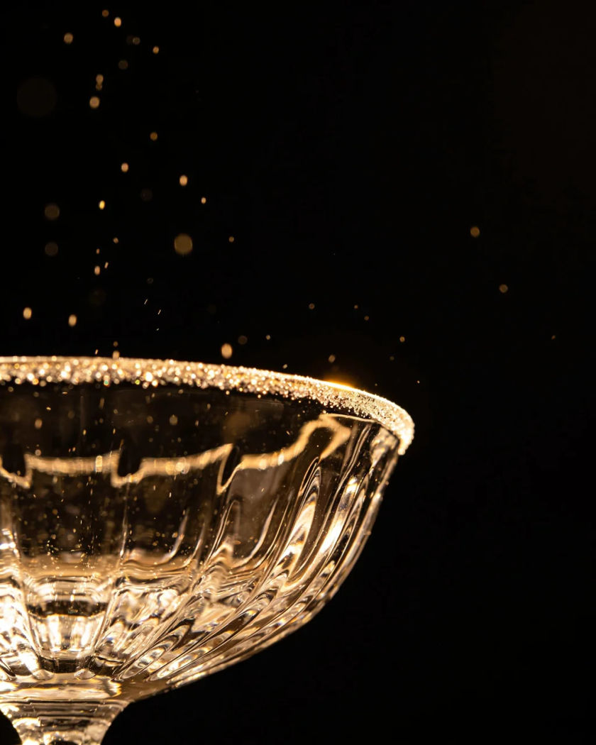 A clear crystal bowl ringing under warm side light against a black ground, fine glowing motes rising from its rim