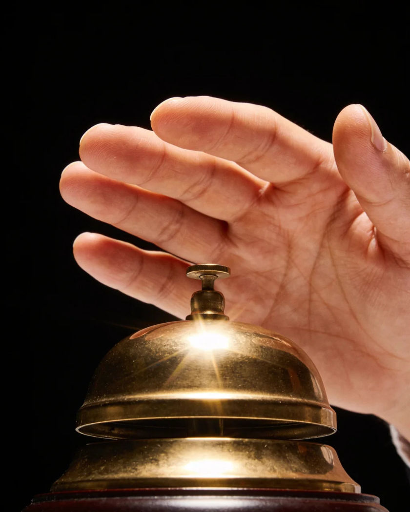 A polished brass desk bell with a palm just lifting off it, a bright glint on the dome under warm light against a pure black ground