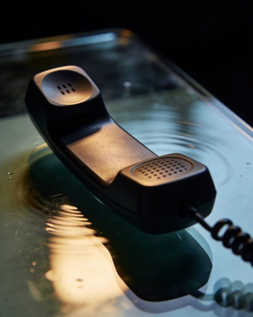 A plain dark handset face down on a glass table, buzzing and shifting under warm side light against a cool dark ground