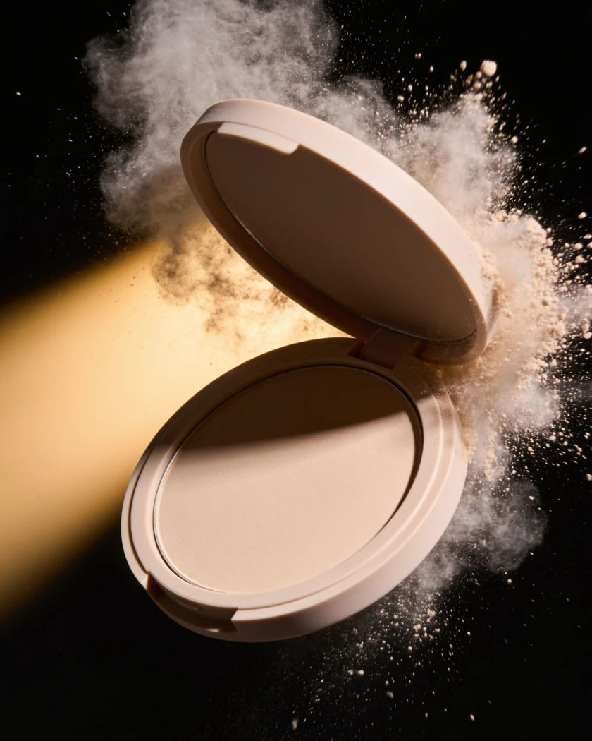A plain round compact snapping shut under warm side light against a black ground, a puff of pale powder bursting from its edge