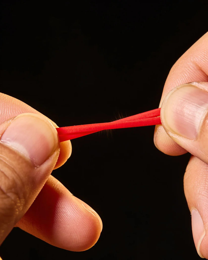 A thick red rubber band pulled taut between two fingers under warm side light against a near-black ground, the band thinning at the point of snapping