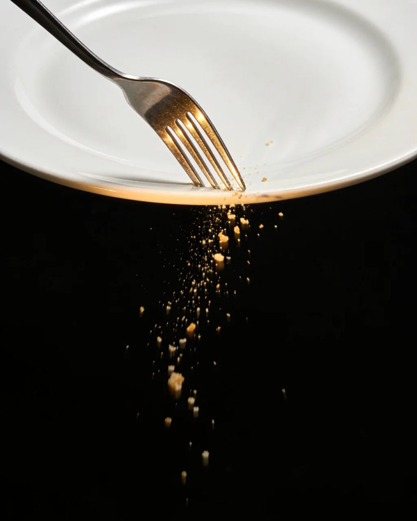 A steel fork striking the rim of a white plate in warm side light against a pure black ground, a tiny spray of crumbs leaping from the impact