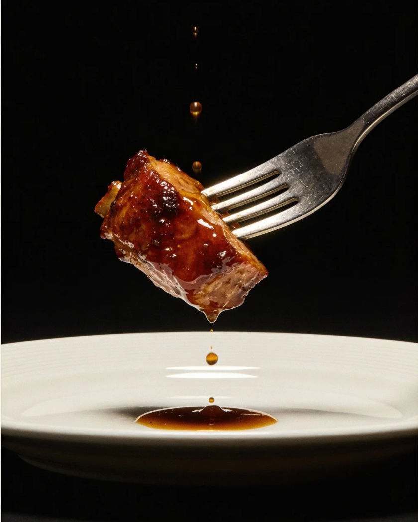 A steel fork lifting a forkful of food off a white plate in warm side light against a dark ground, sauce droplets falling back to the plate
