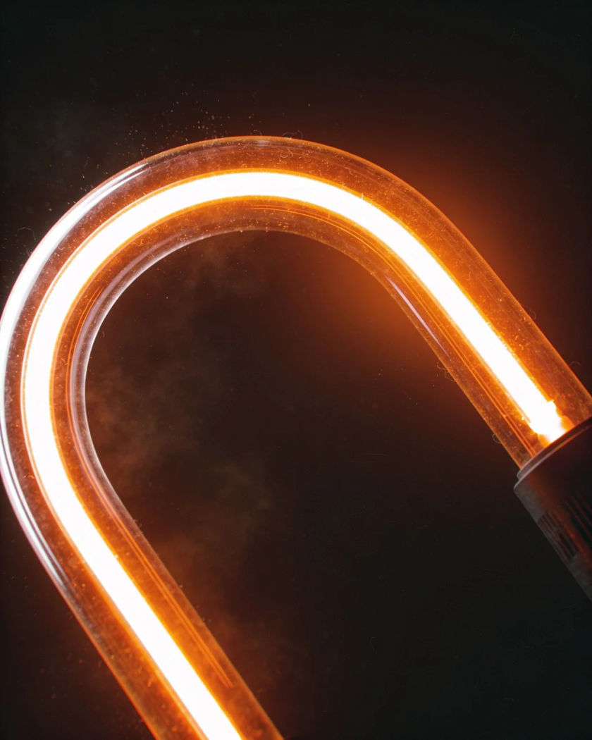 A bent glass neon tube glowing warm orange in close-up against a dark ground, a faint haze of light around the electrode cap
