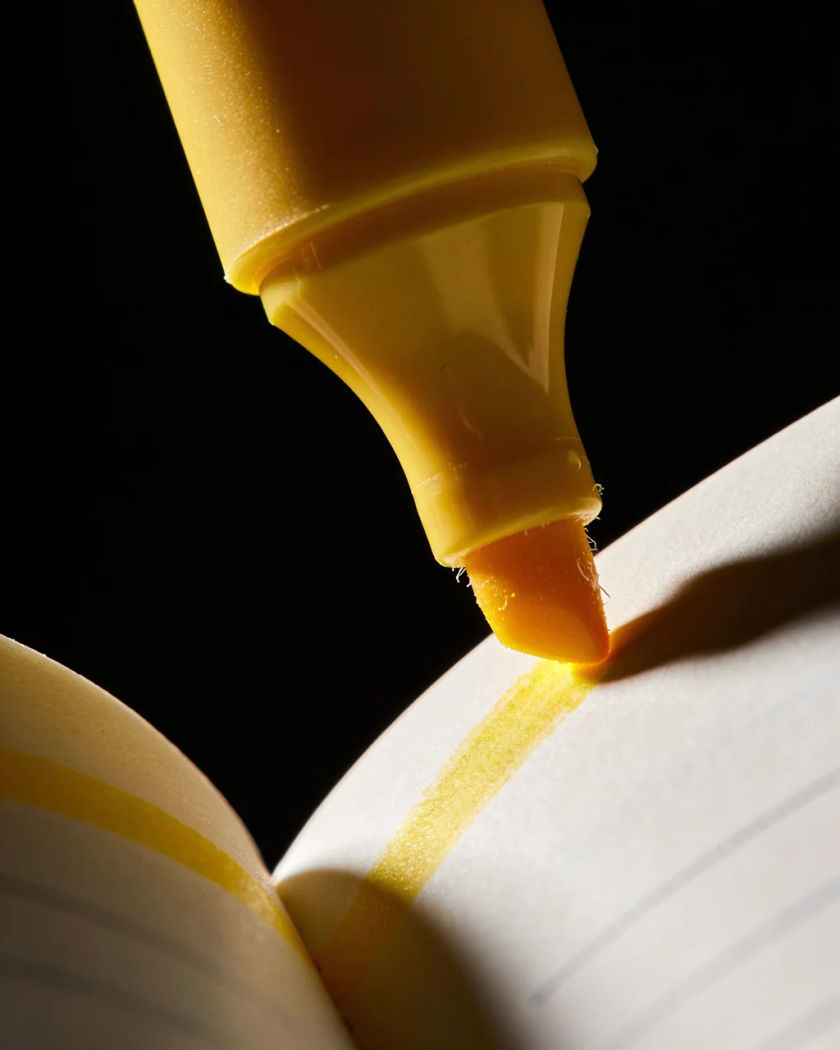 A plain unbranded yellow highlighter dragging a bright stroke across a white page under warm side light against a dark ground, the wet ink glistening