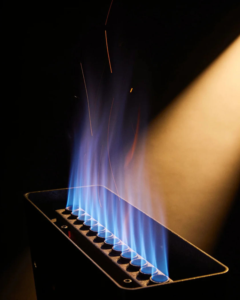 A plain metal furnace with its burner flames glowing blue through an open panel under warm side light against a dark ground