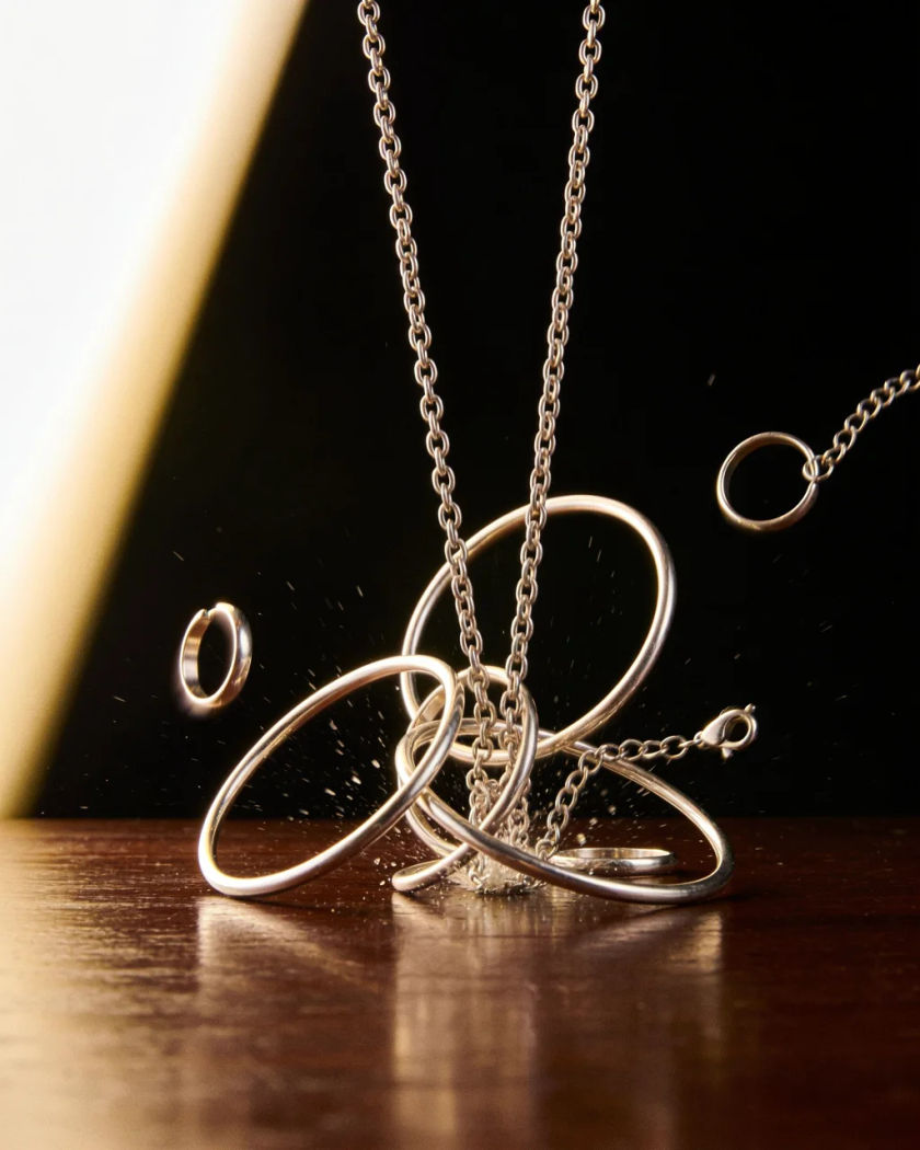 A tangle of plain metal necklaces and bangles crashing onto a wooden table under warm side light against a dark ground, rings bouncing in the air