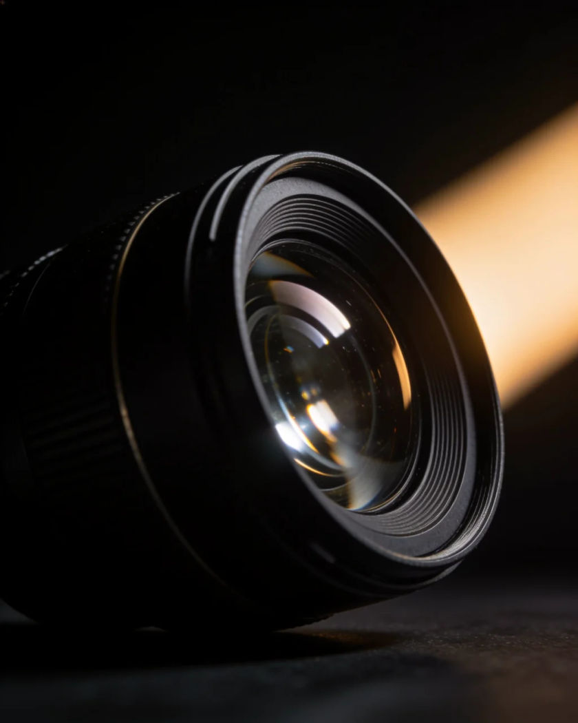 A smooth matte black lens barrel extending under warm side light against a black ground, glints sliding across the glass