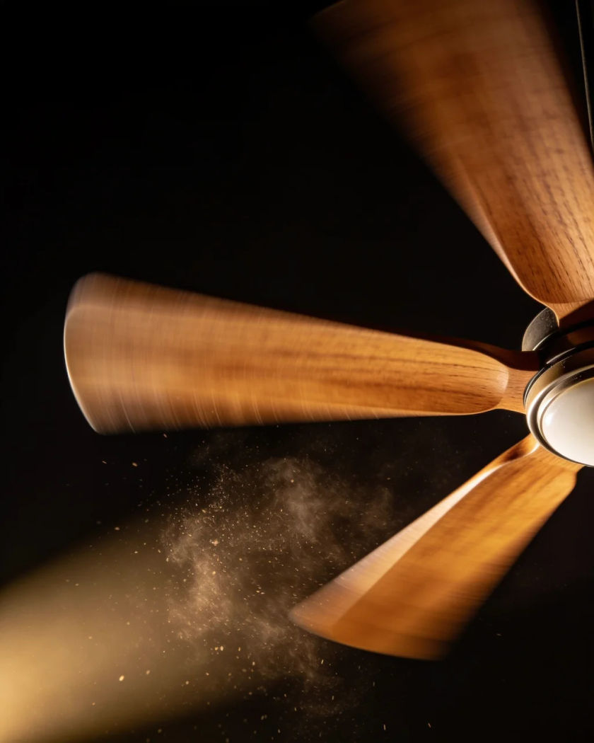 A wooden-bladed ceiling fan spinning, seen from below under warm light against a black ground, dust swirling beneath it