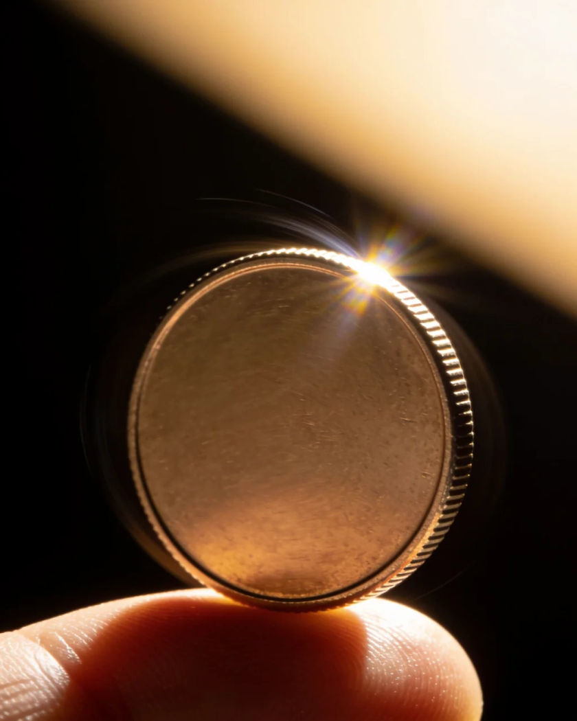 A coin spinning in mid-air above a thumb under warm side light against a black ground, a bright glint flaring off its edge