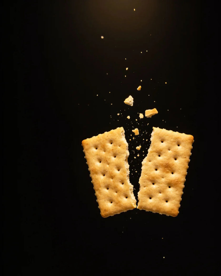 A square cracker snapping in two under warm overhead light against a black ground, crumbs flying from the break