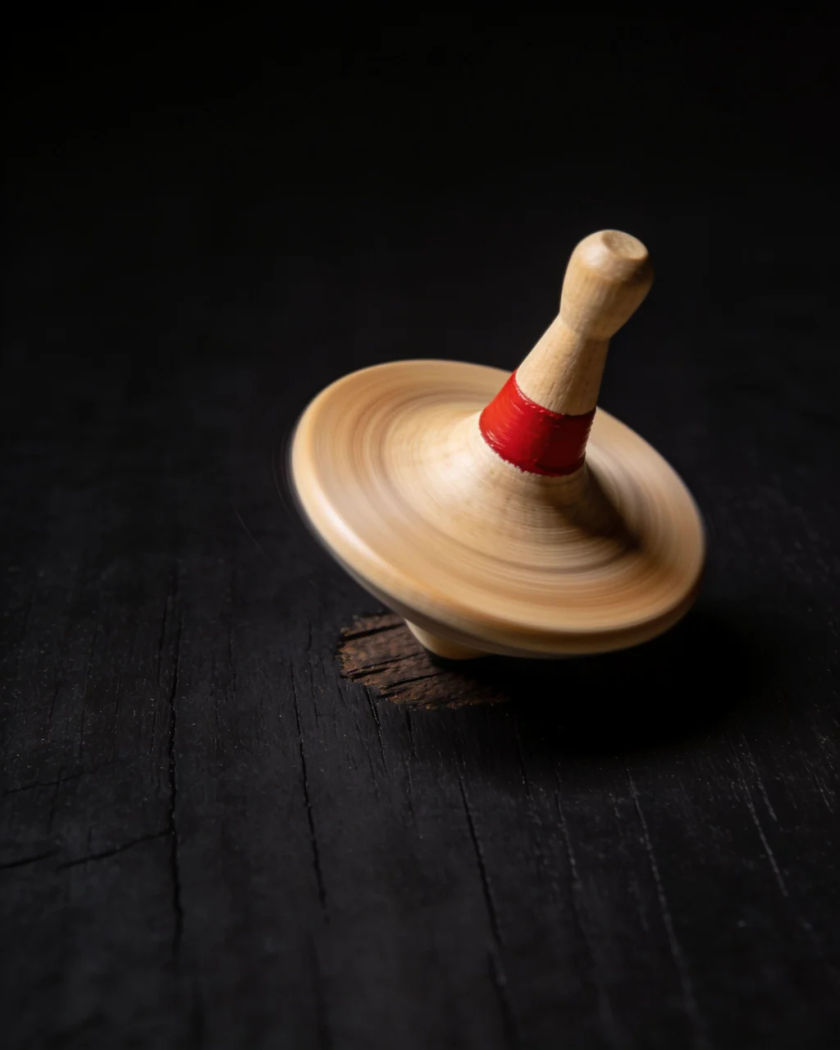 A metal spinning top tilting as it slows on a wooden table under one warm light from above, its blur fading into sharp edges