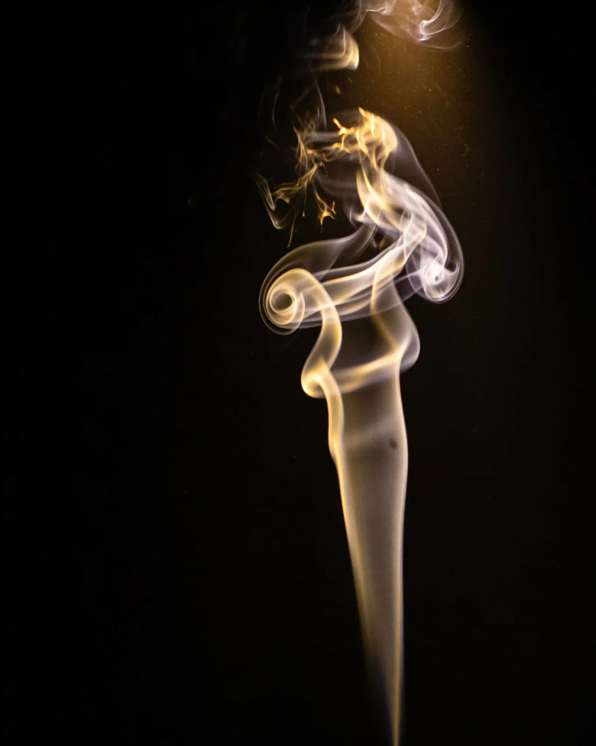 A plume of pale glowing smoke dissolving into the dark under warm light from above against a black ground