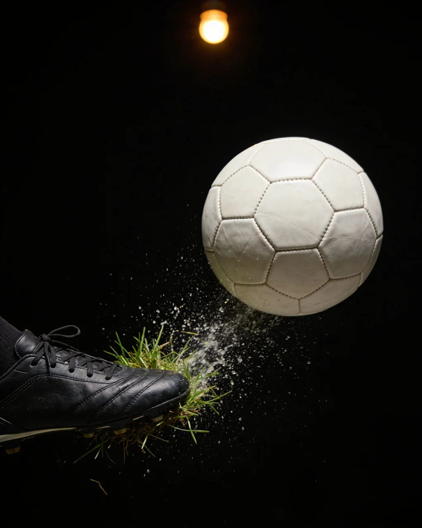 A leather football leaving a boot on wet grass under one warm light from above, turf and water droplets spraying off the strike