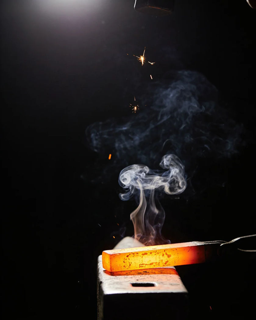 A glowing iron bar plunging into water under warm overhead light against a black ground, white steam bursting up
