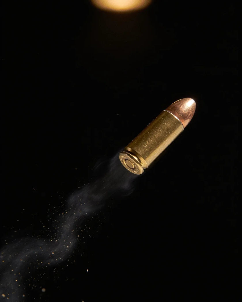 A single brass bullet suspended in mid-air under warm overhead light on black, a thin ripple of air trailing behind it