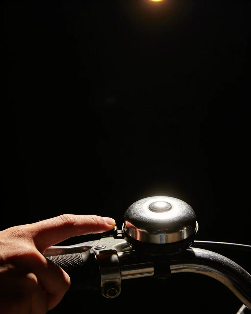A round chrome bicycle bell caught mid-ring under warm light from above against a black ground, a thumb pressing its lever