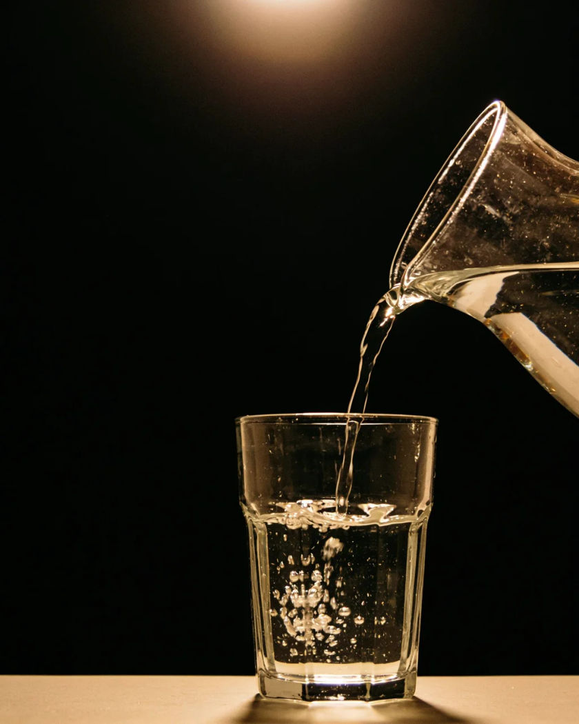 A clear glass jug pouring water into a plain tumbler under warm light from above against a black ground, the stream glinting