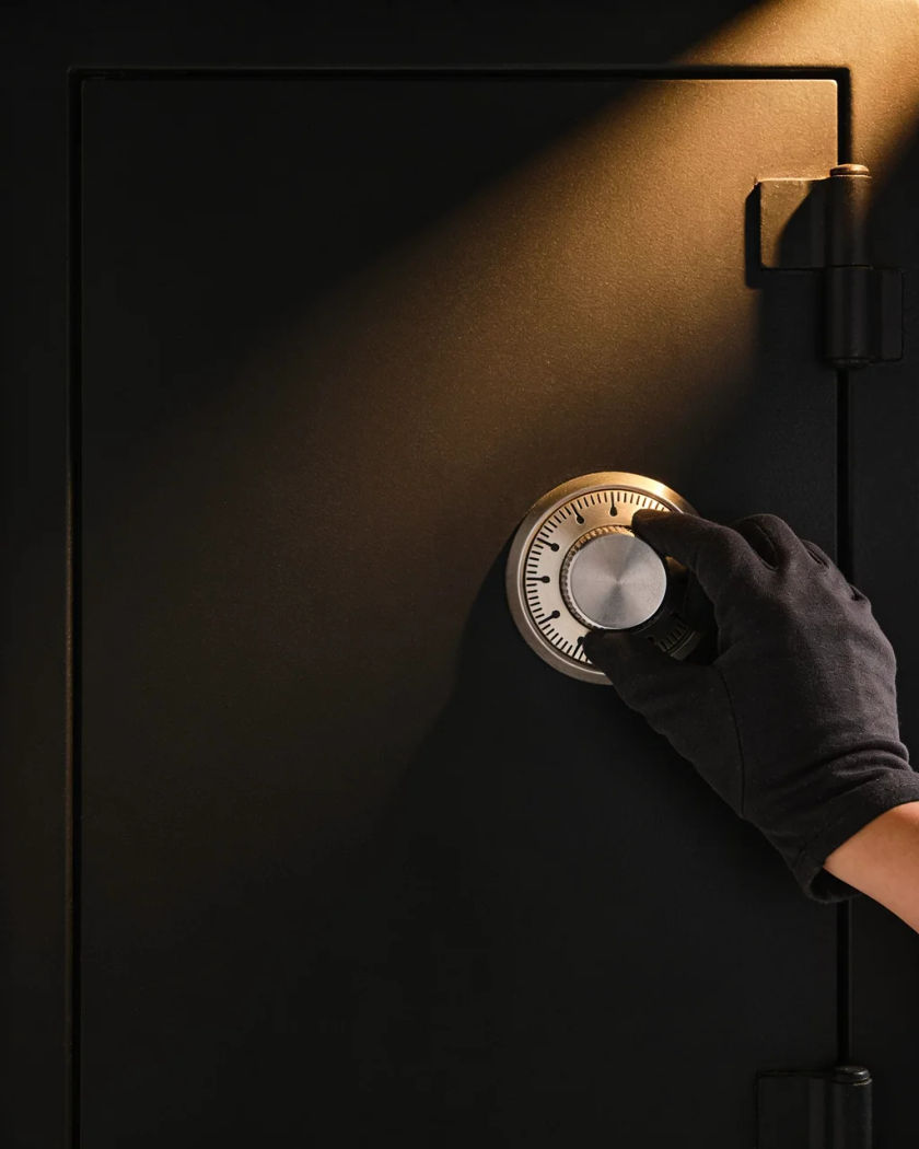 A plain steel safe dial lit by a narrow warm beam on black, a gloved hand turning it