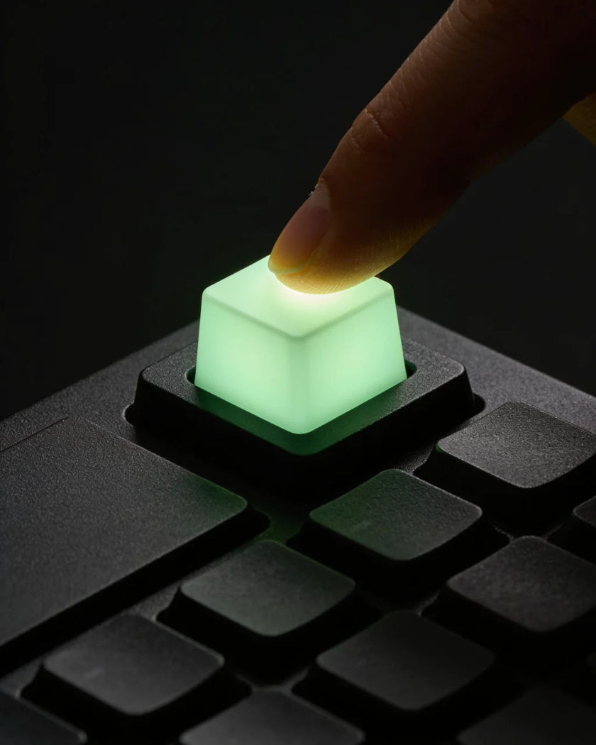 A fingertip resting on one plain blank square key glowing pale green on a dark matte keypad, the key light spilling onto the fingertip