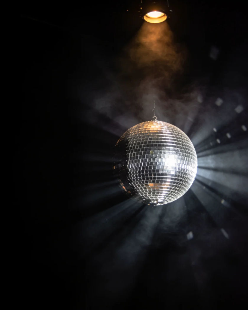 A mirror ball turning under warm light from above against a black ground, small tiles throwing bright beams through haze