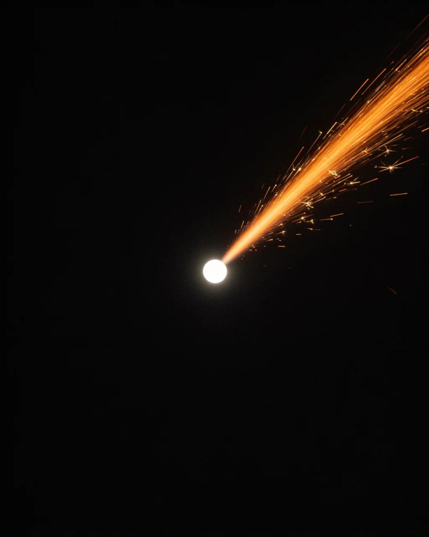 A small glowing orb of white light streaking upward through darkness, a long tail of orange motion blur and fine sparks trailing behind it