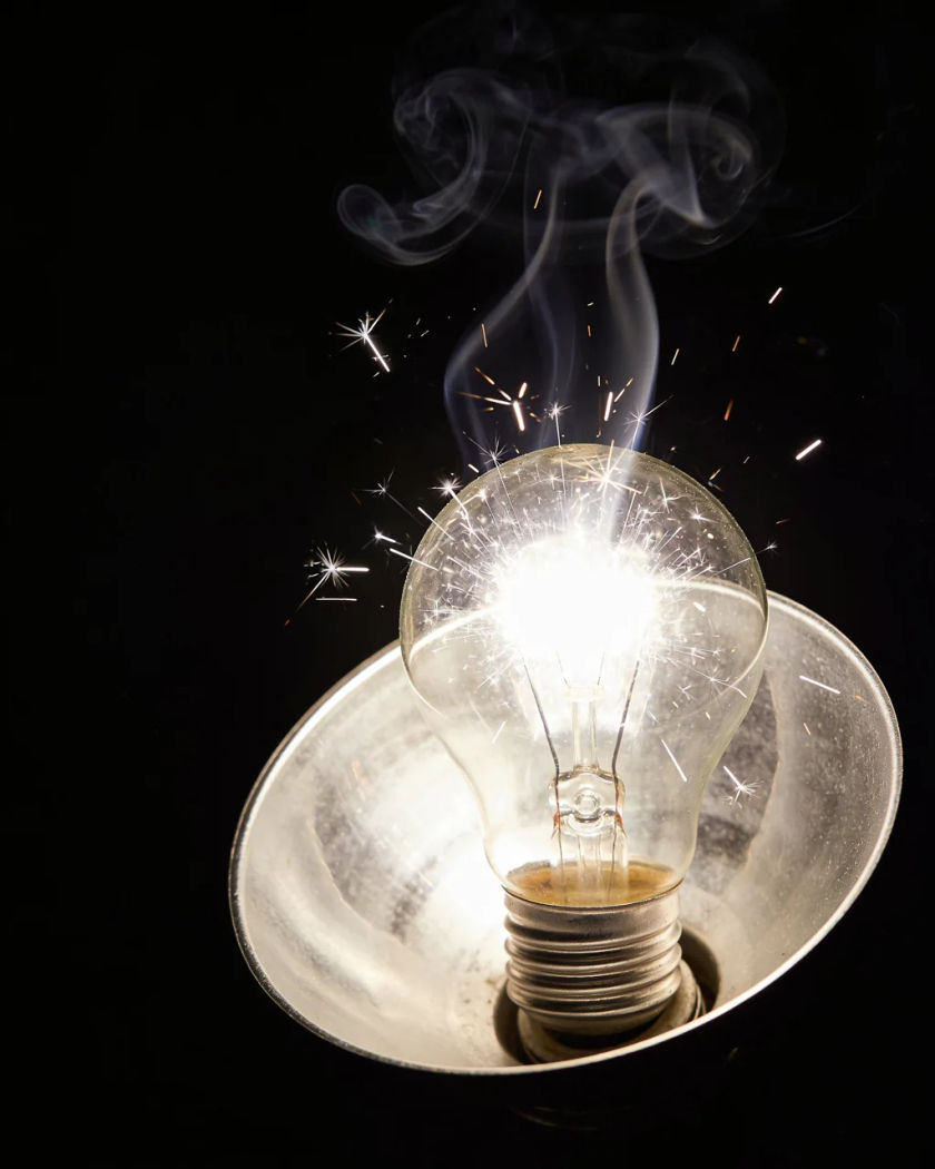 A clear glass flashbulb bursting with white light inside a silver reflector dish, fine sparks and smoke curling out of the glare