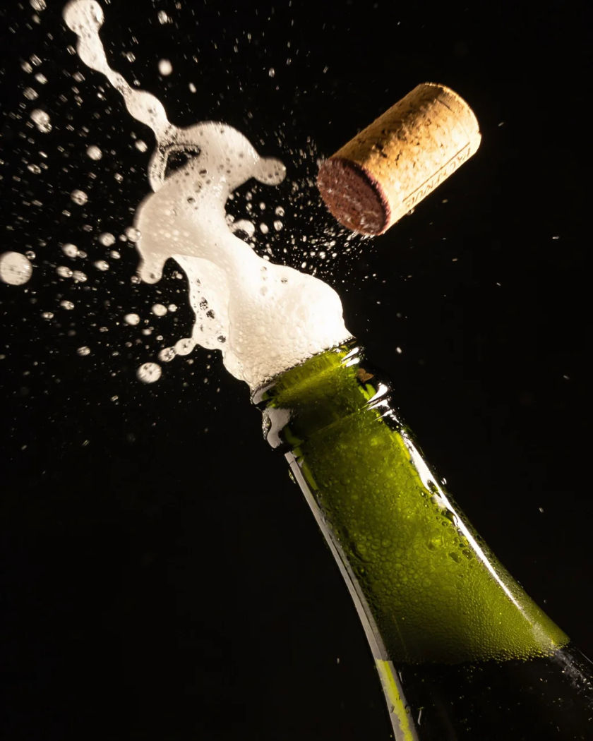 A dark green champagne bottle as its cork flies out, white foam and bright droplets spraying through warm light