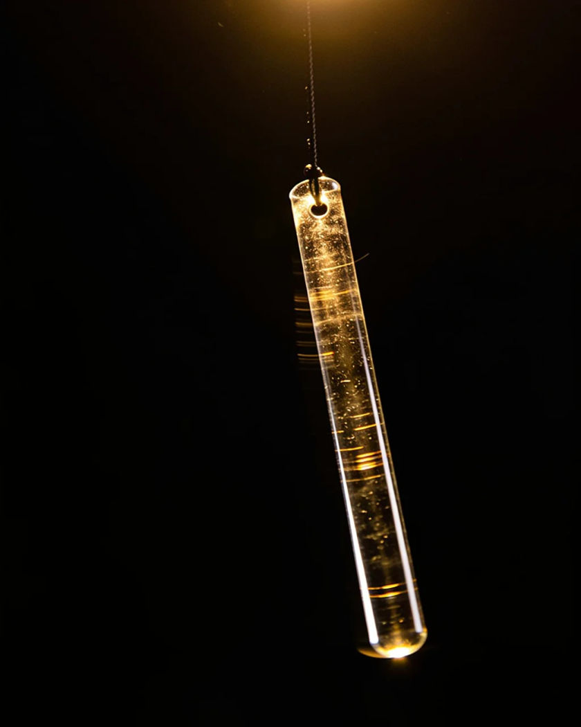 Short clear glass wind chime rods caught mid-sway under warm light from above against a black ground, glints flashing along the glass