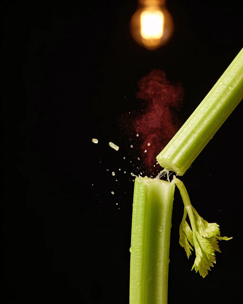 A green celery stalk snapping in half under warm overhead light on black, wet fragments and droplets flying