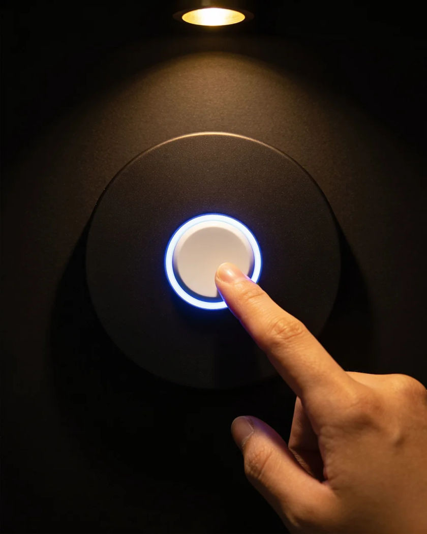 A plain round doorbell button glowing soft blue on black, a fingertip about to press it under warm light from above