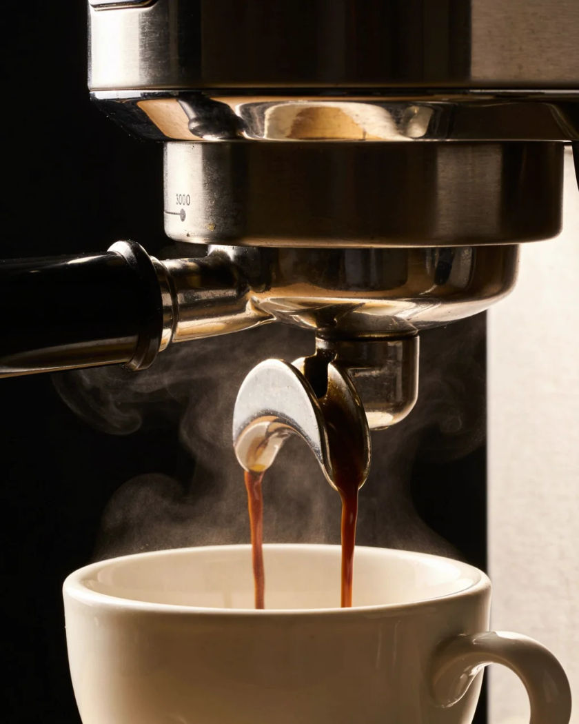 A chrome espresso spout pouring a dark stream of coffee into a small white cup, steam curling up in warm light