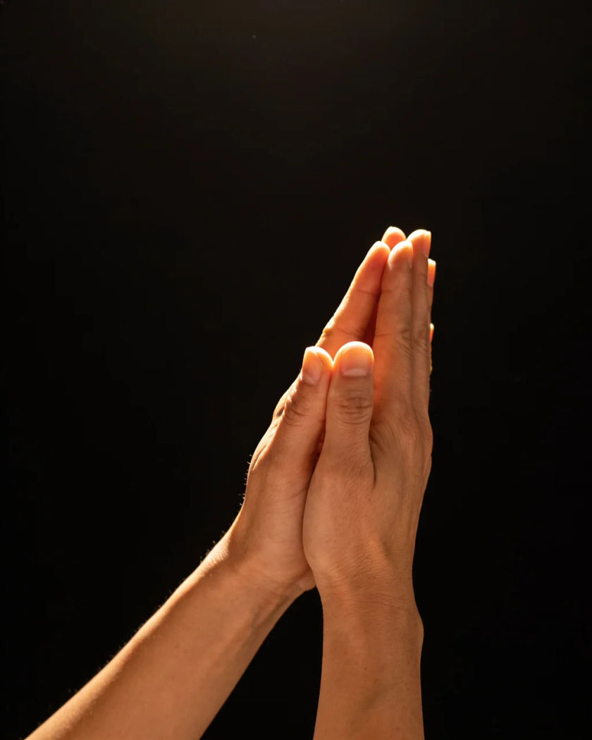 Two adult hands with fingertips pressed together in a steeple, lit from above against a pure black ground