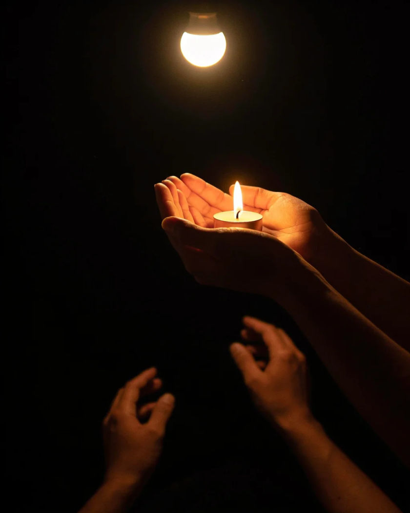 Raised arms of a crowd swaying from behind with small points of warm light under a single overhead light against a black background