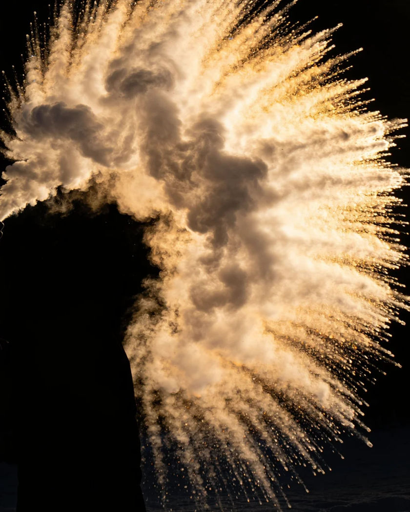 A plume of breath vapour bursting outward in cold air with droplets catching warm light against a black background