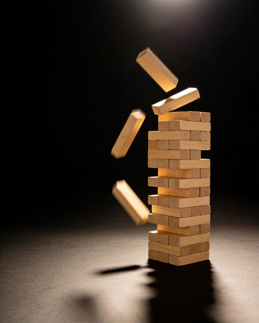 A tower of plain wooden blocks tipping over mid fall under warm light against a black background
