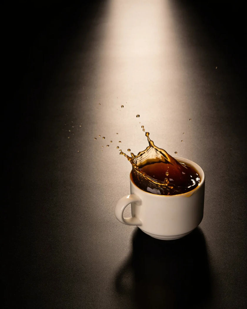 A plain white coffee cup set down hard with a splash of coffee leaping out and droplets frozen mid-air, warm light against a near-black ground
