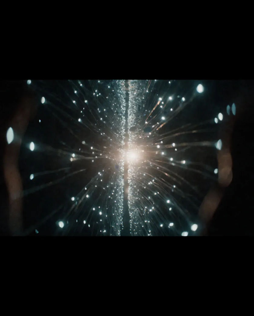 Scattered points of white light streaming toward the center of a black frame and converging into a bright vertical flare