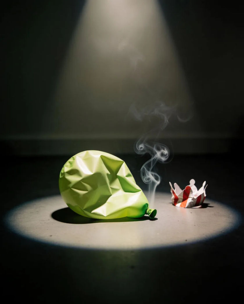 A single deflated party balloon lying on a bare black floor next to a small crumpled paper crown, lit by one overhead spot